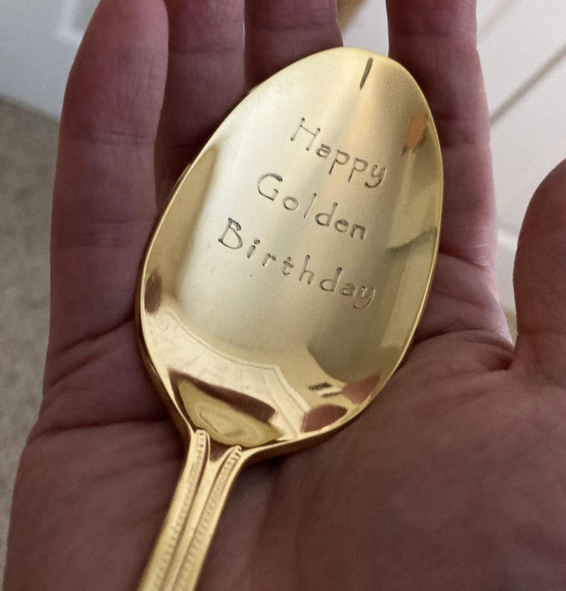 May include: A gold-colored spoon with the words "Happy Golden Birthday" engraved on the bowl. The spoon has a decorative handle and is held in a hand.