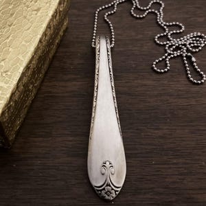 Puede incluir: Un collar de cuchara de plata con un dise&ntilde;o floral decorativo en el extremo. La cuchara est&aacute; unida a una cadena de bolas de plata.