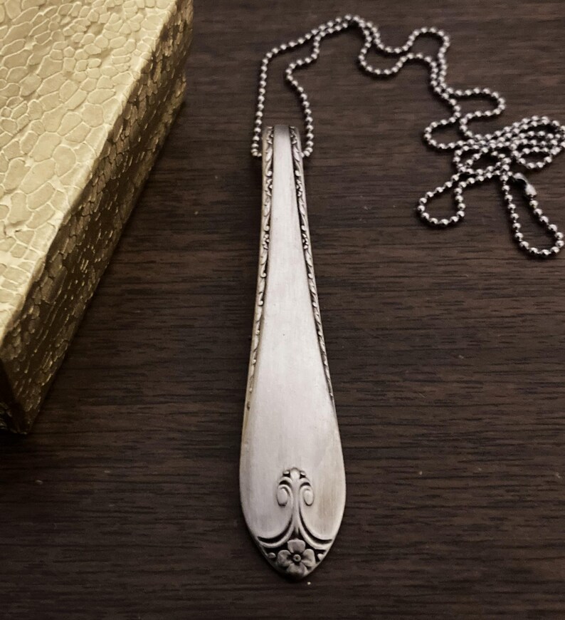 Puede incluir: Un collar de cuchara de plata con un dise&ntilde;o floral decorativo en el extremo. La cuchara est&aacute; unida a una cadena de bolas de plata.