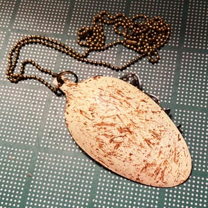 May include: A white spoon with brown paint splatters, attached to a gold chain necklace. The spoon is shaped like a teardrop and has a small loop at the top.