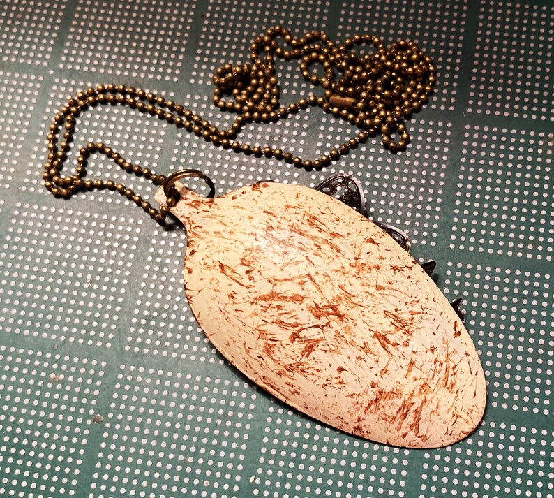 May include: A white spoon with brown paint splatters, attached to a gold chain necklace. The spoon is shaped like a teardrop and has a small loop at the top.