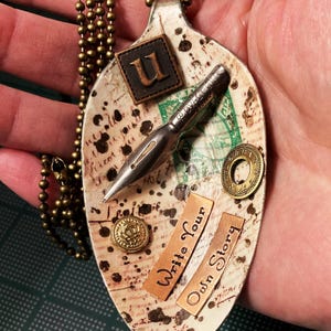 Op de afbeelding: Een witte lepelsnoer met een vintage uitstraling. De lepel is versierd met een pen, een postzegel, een knoop en twee koperen labels met de tekst "Write Your Own Story". De lepel is bevestigd aan een gouden ketting.