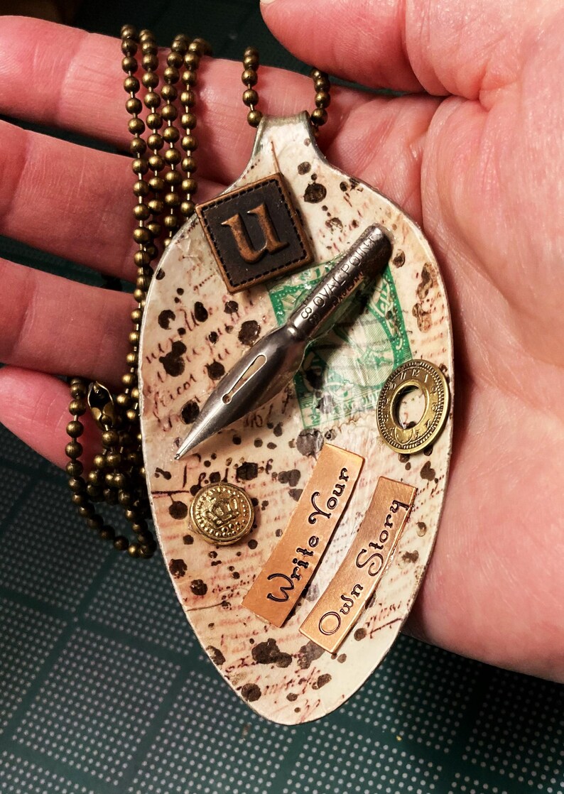 Op de afbeelding: Een witte lepelsnoer met een vintage uitstraling. De lepel is versierd met een pen, een postzegel, een knoop en twee koperen labels met de tekst "Write Your Own Story". De lepel is bevestigd aan een gouden ketting.