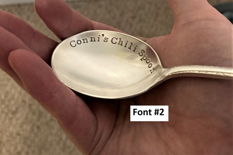 May include: A silver spoon with the text "Conni's Chili Spoon" engraved on the bowl. The spoon is being held in a hand.