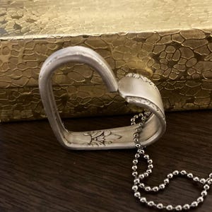 Puede incluir: Un collar con un colgante en forma de coraz&oacute;n de plata hecho de una cuchara reutilizada. El colgante tiene un dise&ntilde;o floral y est&aacute; unido a una cadena de plata.