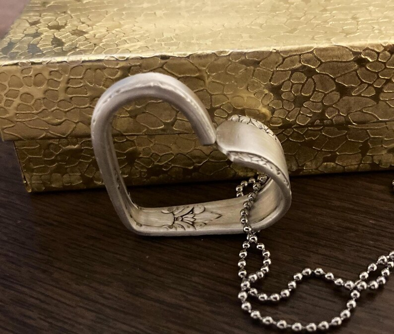 Puede incluir: Un collar con un colgante en forma de coraz&oacute;n de plata hecho de una cuchara reutilizada. El colgante tiene un dise&ntilde;o floral y est&aacute; unido a una cadena de plata.