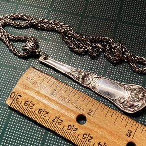 May include: A silver spoon necklace with a detailed floral design. The spoon is attached to a silver chain. The spoon is about 7 inches long. The chain is about 18 inches long.