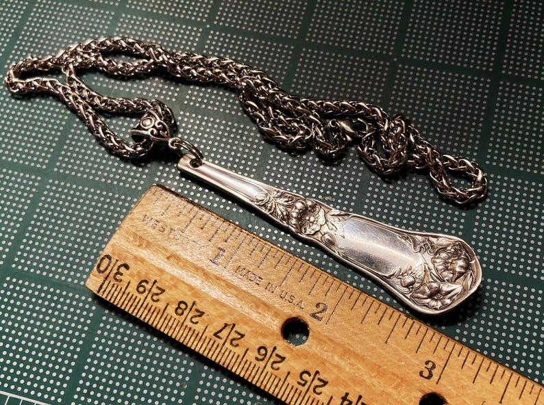 May include: A silver spoon necklace with a detailed floral design. The spoon is attached to a silver chain. The spoon is about 7 inches long. The chain is about 18 inches long.