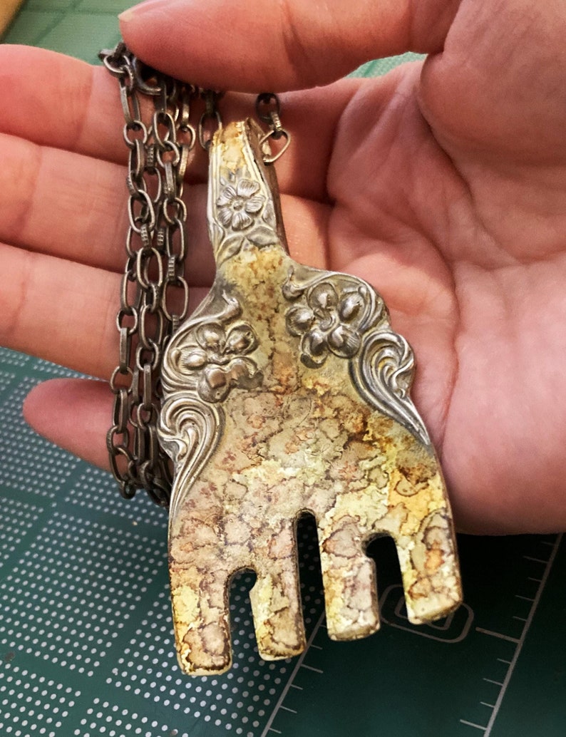May include: A silver fork necklace with a tarnished finish and ornate floral details. The fork is attached to a silver chain.
