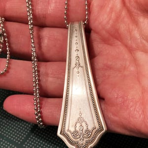 May include: A silver spoon necklace with a decorative design on the handle. The spoon is attached to a silver ball chain.