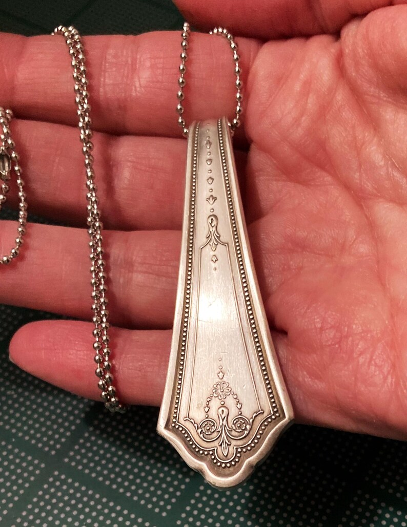 May include: A silver spoon necklace with a decorative design on the handle. The spoon is attached to a silver ball chain.