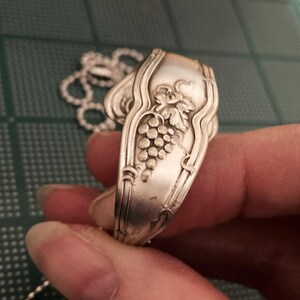 May include: A silver spoon necklace with a grape design. The spoon is bent into a circle and has a chain attached to it.