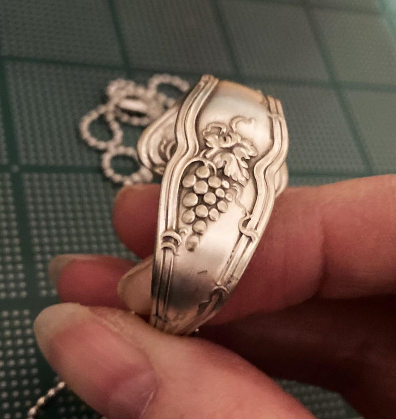 May include: A silver spoon necklace with a grape design. The spoon is bent into a circle and has a chain attached to it.