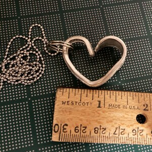 May include: A silver heart-shaped pendant necklace with a ball chain. The pendant is about 1 inch wide and is made from a recycled metal. The necklace is shown next to a ruler.