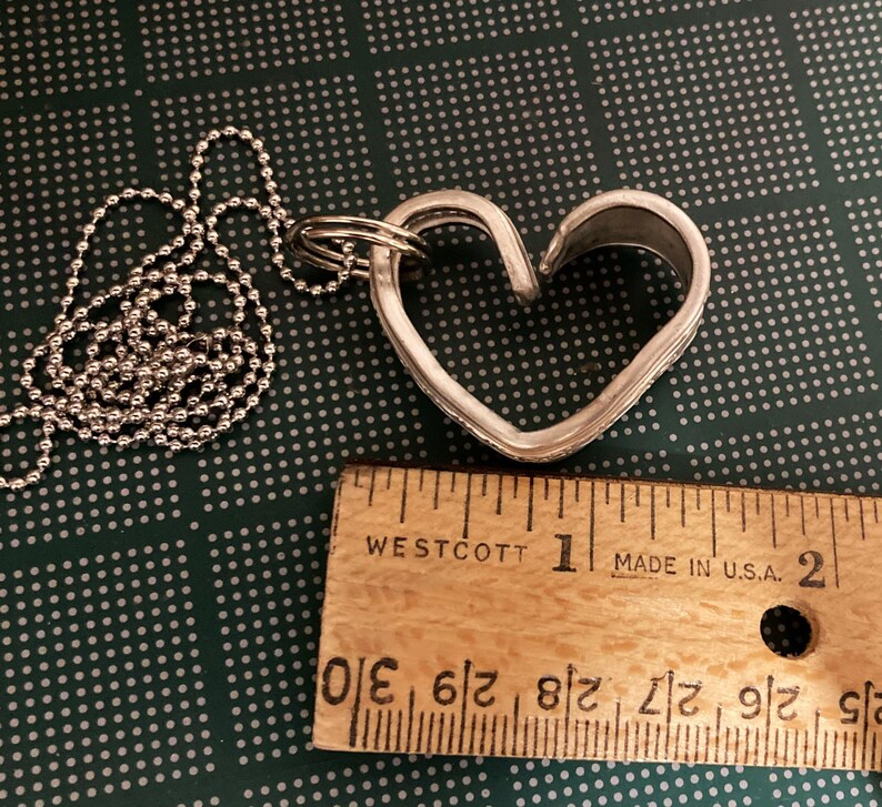 May include: A silver heart-shaped pendant necklace with a ball chain. The pendant is about 1 inch wide and is made from a recycled metal. The necklace is shown next to a ruler.