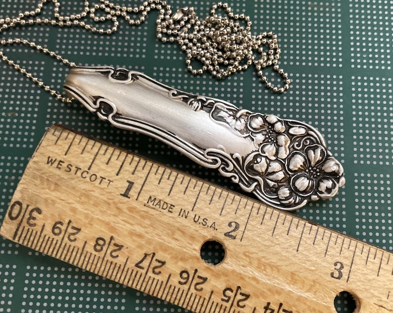 May include: A silver spoon necklace with a floral design. The spoon is approximately 2 inches long. The necklace is made with a silver chain.