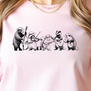 May include: Light pink t-shirt featuring a black and white illustration of a band of animals playing musical instruments. The band includes a bear, raccoon, and other animals. The shirt has the text "SERAPHINE WEAR PERSONALIZED GIFTS" on the upper left.