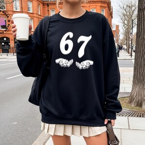 May include: Black sweatshirt with the number "67" in white script and two white hand graphics. The sweatshirt is paired with a cream-colored pleated skirt and white knee-high socks. A black shoulder bag and a brown purse are also visible.