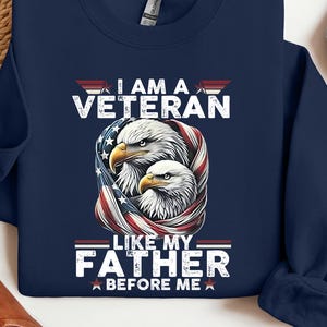 May include: Navy blue sweatshirt with the text "I AM A VETERAN LIKE MY FATHER BEFORE ME." The design features two eagles wrapped in an American flag. The sweatshirt is a crew neck style.