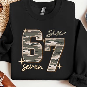 May include: Black sweatshirt with the numbers "67" in a camouflage pattern, with the words "six" and "seven" in a cursive font. The design includes star accents. The sweatshirt is paired with blue jeans and brown shoes.