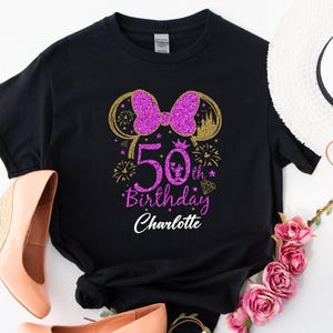 Puede incluir: Camiseta negra con un lazo morado brillante y un dise&ntilde;o de orejas doradas, celebrando un 50 cumplea&ntilde;os. La camiseta incluye el texto "50th Birthday Charlotte". Zapatos de tac&oacute;n cu&ntilde;a color melocot&oacute;n y un sombrero blanco con detalles florales.