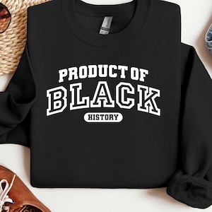 K&ouml;nnte beinhalten: Schwarzes Sweatshirt mit dem wei&szlig;en Text "PRODUCT OF BLACK HISTORY" im College-Stil. Das Bild zeigt auch braune Schuhe und blaue Jeans, was auf einen l&auml;ssigen Stil hindeutet.