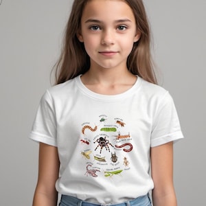 May include: White t-shirt featuring a colorful insect graphic. The design includes illustrations of various bugs, such as a spider, scorpion, and grasshopper, with their names. The shirt is worn by a young person.