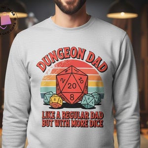May include: Light gray sweatshirt with the text "DUNGEON DAD" above a graphic of a red twenty-sided die and other dice. The text "LIKE A REGULAR DAD BUT WITH MORE DICE" is below the graphic. The design is in a retro style.