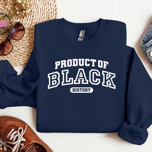 K&ouml;nnte beinhalten: Marineblaues Sweatshirt mit dem wei&szlig;en Text "PRODUCT OF BLACK HISTORY" in einem College-Stil. Das Bild zeigt braune Schuhe, eine Sonnenbrille und eine Jeans. Das Sweatshirt ist gefaltet.