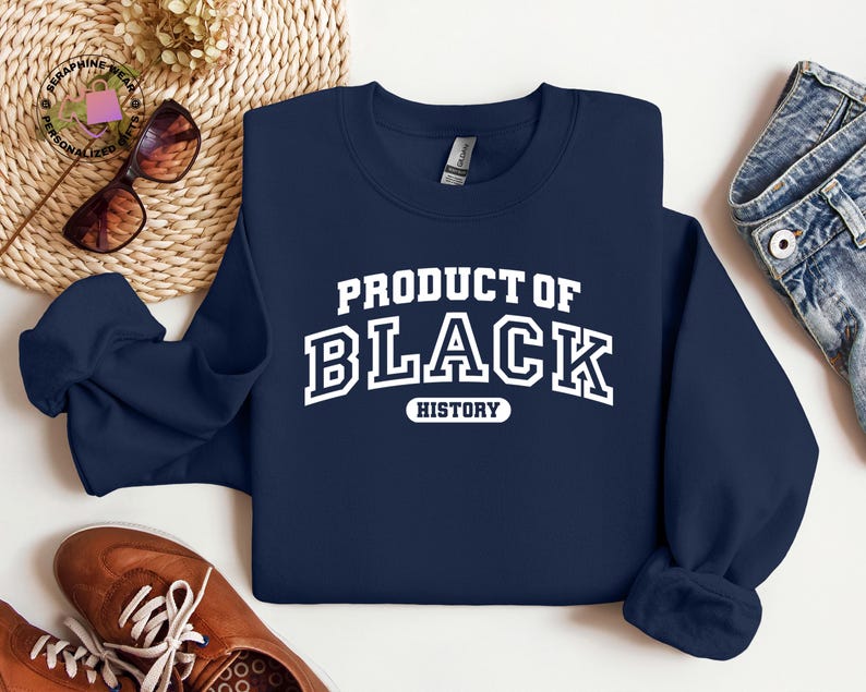 K&ouml;nnte beinhalten: Marineblaues Sweatshirt mit dem wei&szlig;en Text "PRODUCT OF BLACK HISTORY" in einem College-Stil. Das Bild zeigt braune Schuhe, eine Sonnenbrille und eine Jeans. Das Sweatshirt ist gefaltet.