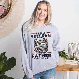 May include: A light grey crew neck sweatshirt with the text "I AM A VETERAN LIKE MY FATHER BEFORE ME" and an eagle graphic. The design incorporates the American flag.