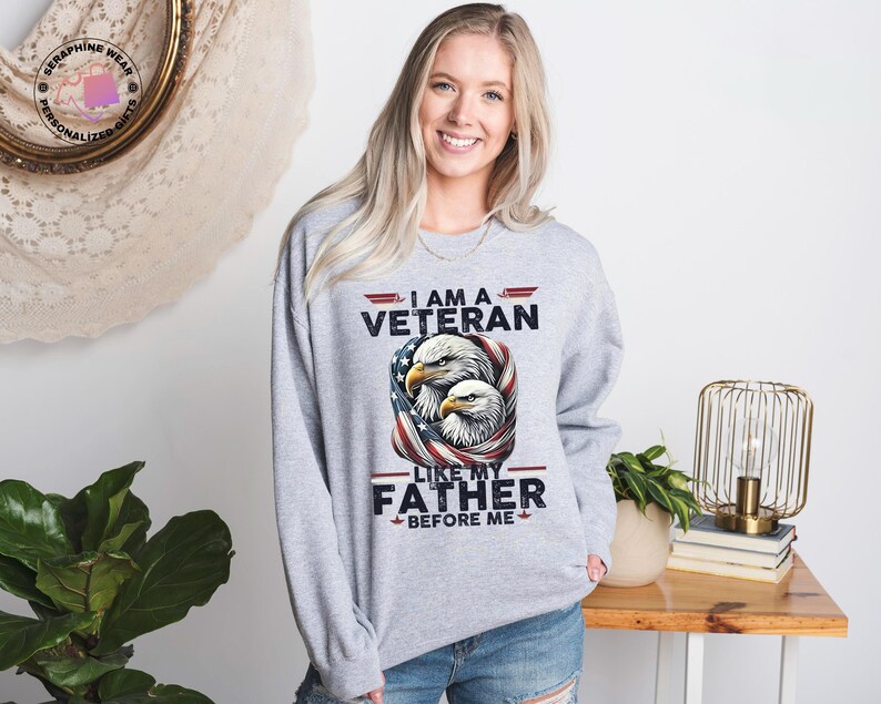May include: A light grey crew neck sweatshirt with the text "I AM A VETERAN LIKE MY FATHER BEFORE ME" and an eagle graphic. The design incorporates the American flag.