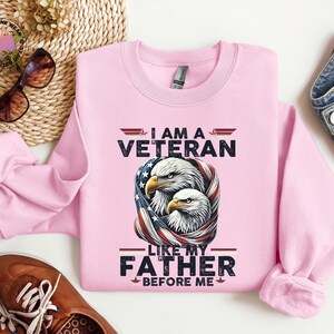 May include: A pale pink sweatshirt featuring the text "I AM A VETERAN LIKE MY FATHER BEFORE ME" and a graphic of two eagles within an American flag design. The sweatshirt is made of a soft fabric.
