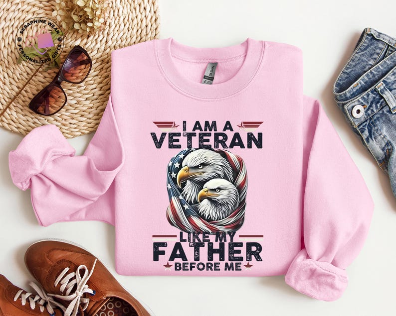 May include: A pale pink sweatshirt featuring the text "I AM A VETERAN LIKE MY FATHER BEFORE ME" and a graphic of two eagles within an American flag design. The sweatshirt is made of a soft fabric.
