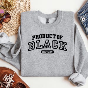 K&ouml;nnte beinhalten: Ein hellgraues Sweatshirt mit dem Text "PRODUCT OF BLACK HISTORY" in schwarzen Buchstaben. Das Bild zeigt braune Schuhe, eine Sonnenbrille und Jeans.