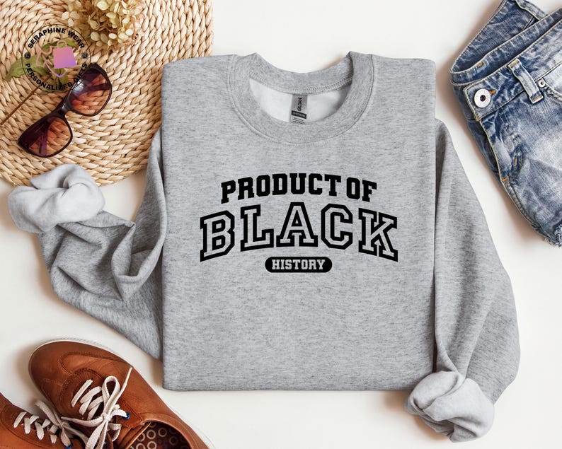 K&ouml;nnte beinhalten: Ein hellgraues Sweatshirt mit dem Text "PRODUCT OF BLACK HISTORY" in schwarzen Buchstaben. Das Bild zeigt braune Schuhe, eine Sonnenbrille und Jeans.