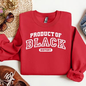 K&ouml;nnte beinhalten: Ein rotes Sweatshirt mit dem wei&szlig;en Schriftzug "PRODUCT OF BLACK HISTORY". Das Bild zeigt Sonnenbrillen, eine gewebte Matte, blaue Jeans und braune Schuhe.