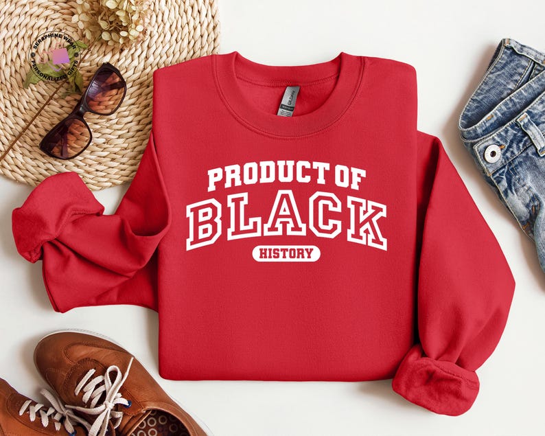K&ouml;nnte beinhalten: Ein rotes Sweatshirt mit dem wei&szlig;en Schriftzug "PRODUCT OF BLACK HISTORY". Das Bild zeigt Sonnenbrillen, eine gewebte Matte, blaue Jeans und braune Schuhe.