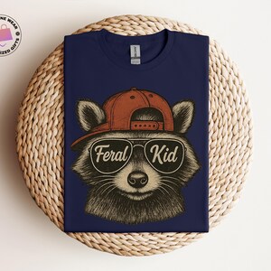 May include: Navy blue t-shirt featuring a raccoon wearing a red baseball cap and sunglasses. The sunglasses have the words "Feral Kid" printed on them. The shirt is a Gildan brand.