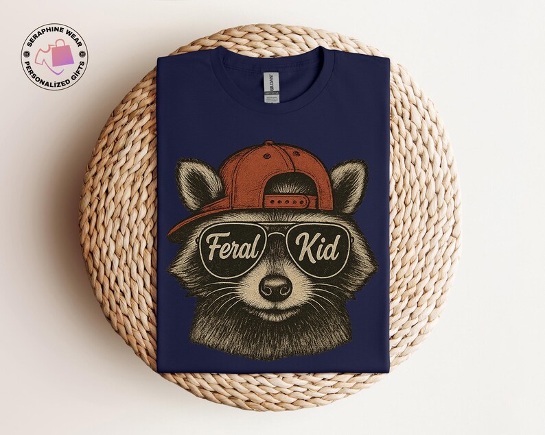 May include: Navy blue t-shirt featuring a raccoon wearing a red baseball cap and sunglasses. The sunglasses have the words "Feral Kid" printed on them. The shirt is a Gildan brand.