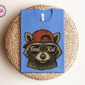May include: A blue t-shirt featuring a raccoon wearing a brown baseball cap and sunglasses. The sunglasses have the words "Feral Kid" printed on them. The shirt is folded on a woven, circular surface.