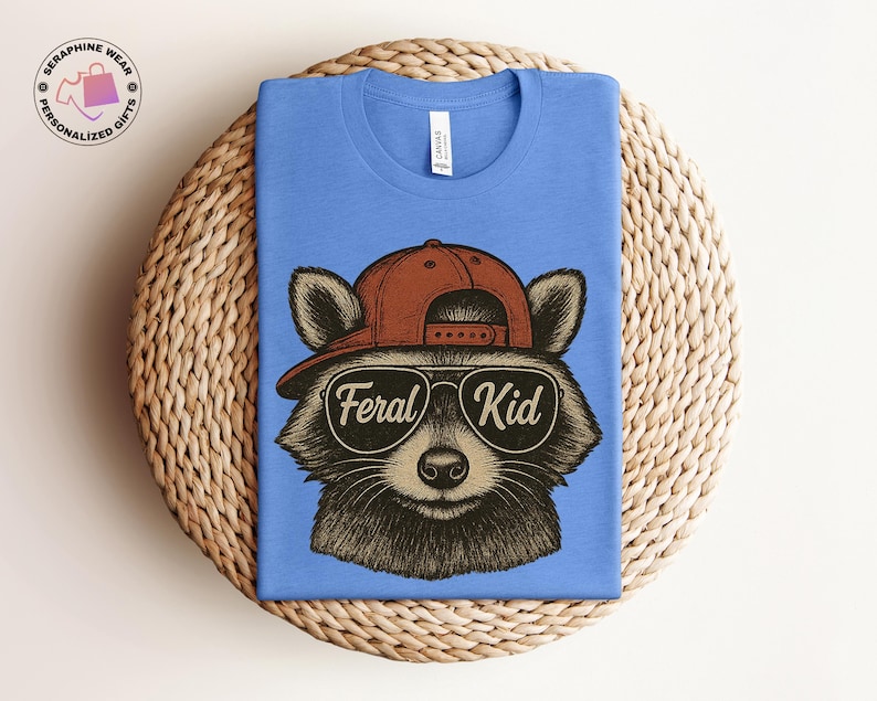 May include: A blue t-shirt featuring a raccoon wearing a brown baseball cap and sunglasses. The sunglasses have the words "Feral Kid" printed on them. The shirt is folded on a woven, circular surface.