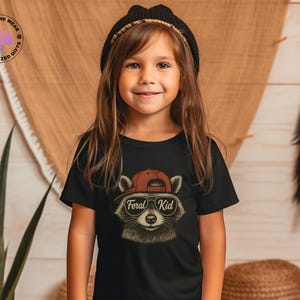 May include: A young girl wearing a black t-shirt with a raccoon graphic. The raccoon is wearing a red cap and sunglasses, with the text "Feral Kid" below. The girl is also wearing a black beanie.