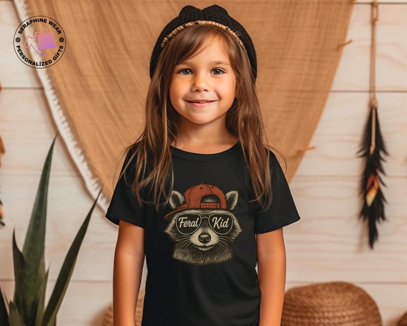 May include: A young girl wearing a black t-shirt with a raccoon graphic. The raccoon is wearing a red cap and sunglasses, with the text "Feral Kid" below. The girl is also wearing a black beanie.
