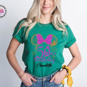 Puede incluir: Camiseta verde esmeralda con un lazo morado brillante y un dise&ntilde;o de orejas de rat&oacute;n doradas. La camiseta presenta el texto "50th Birthday Charlotte" en una fuente cursiva. La camiseta es de cuello redondo y tiene mangas enrolladas.