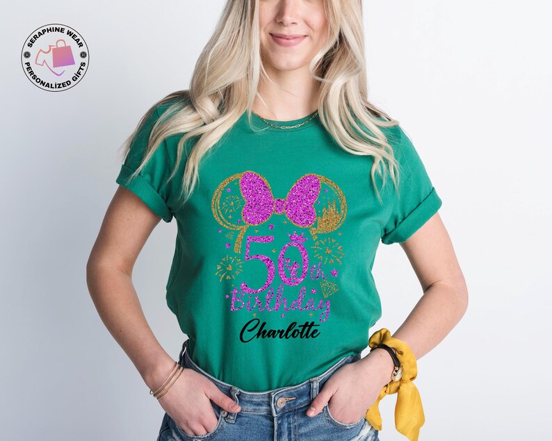 Puede incluir: Camiseta verde esmeralda con un lazo morado brillante y un dise&ntilde;o de orejas de rat&oacute;n doradas. La camiseta presenta el texto "50th Birthday Charlotte" en una fuente cursiva. La camiseta es de cuello redondo y tiene mangas enrolladas.