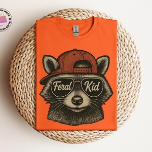 May include: Orange t-shirt featuring a raccoon wearing a brown cap and sunglasses with the text "Feral Kid". The raccoon illustration is in black and white. The shirt is displayed on a woven, circular surface.