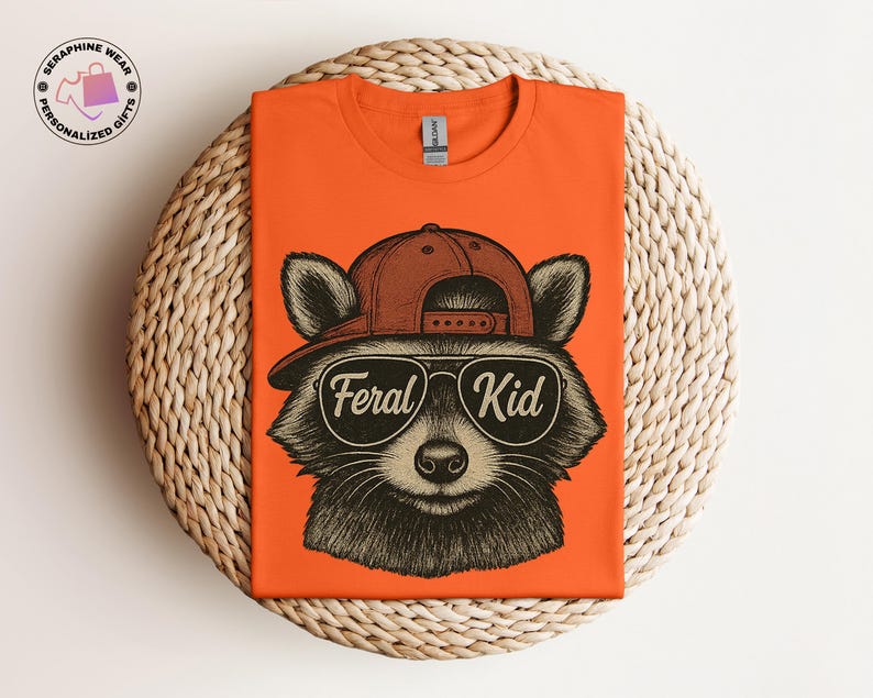 May include: Orange t-shirt featuring a raccoon wearing a brown cap and sunglasses with the text "Feral Kid". The raccoon illustration is in black and white. The shirt is displayed on a woven, circular surface.