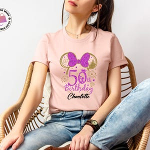 Puede incluir: Camiseta rosa claro con un lazo y orejas de Minnie Mouse morado brillante. La camiseta presenta el texto "50th Birthday Charlotte" en morado y negro, con fuegos artificiales y una corona. Un regalo de cumplea&ntilde;os personalizado.