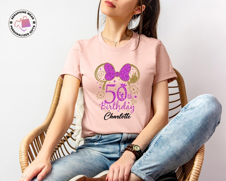 Puede incluir: Camiseta rosa claro con un lazo y orejas de Minnie Mouse morado brillante. La camiseta presenta el texto "50th Birthday Charlotte" en morado y negro, con fuegos artificiales y una corona. Un regalo de cumplea&ntilde;os personalizado.